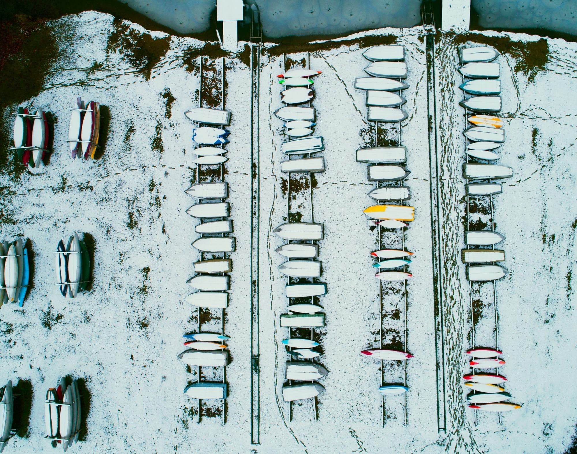 Overhead winter drone photo of small sailboats and dinghies racked on a snow-covered shore
